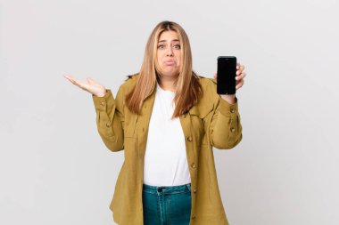 Kıvrımlı, güzel, sarışın kadın kafası karışık, şüpheci ve akıllı bir telefon tutuyor.