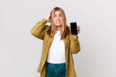 Kıvrımlı, güzel, sarışın kadın stresli, endişeli ya da korkmuş hissediyor, elleri başının üstünde ve akıllı bir telefon tutuyor.