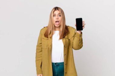 Kıvrımlı, güzel, sarışın kadın şok olmuş ya da şaşırmış görünüyor ve elinde akıllı bir telefon tutuyor.