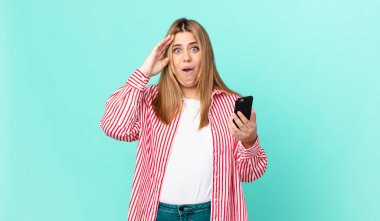 Kıvrımlı, güzel sarışın kadın mutlu, şaşkın ve şaşırmış görünüyor ve elinde akıllı bir telefon tutuyor.