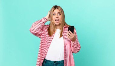 Kıvrımlı, güzel, sarışın kadın stresli, endişeli ya da korkmuş hissediyor, elleri başının üstünde ve akıllı bir telefon tutuyor.