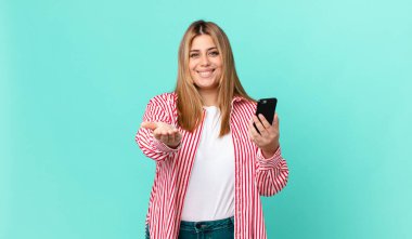 kıvrımlı, güzel sarışın kadın mutlu bir şekilde gülümsüyor ve bir konsept sunuyor ve akıllı bir telefon tutuyor.