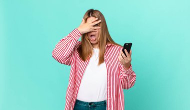 Kıvrımlı, güzel, sarışın kadın şok olmuş, korkmuş ya da korkmuş görünüyor, yüzünü eliyle kapatıyor ve akıllı bir telefon tutuyor.