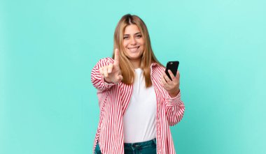 Kıvrımlı, güzel sarışın kadın gururla gülümsüyor ve kendine güvenerek bir numara oluyor ve elinde akıllı bir telefon tutuyor.