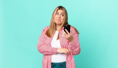 kıvrımlı, güzel, sarışın bir kadın omuz silkiyor, kafası karışık ve kararsız hissediyor ve elinde akıllı bir telefon tutuyor.