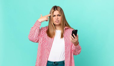 kıvrımlı, güzel sarışın kadın kafası karışık ve şaşkın hissediyor, deli olduğunu ve elinde akıllı bir telefon tuttuğunu gösteriyor.
