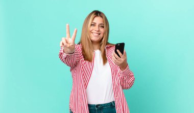 Kıvrımlı, güzel sarışın kadın gülümsüyor ve arkadaş canlısı görünüyor. İki numarayı gösteriyor ve elinde akıllı bir telefon var.