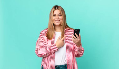 Kıvrımlı, güzel, sarışın bir kadın neşeyle gülümsüyor, mutlu hissediyor ve bir yandan işaret ediyor ve akıllı bir telefon tutuyor.