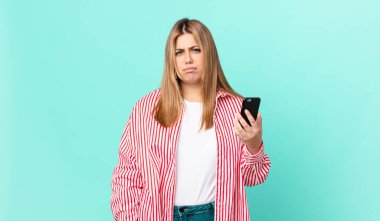 Kıvrımlı, güzel, sarışın bir kadın üzgün, üzgün ya da kızgın hissediyor ve kenara bakıyor ve akıllı bir telefon tutuyor.