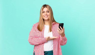 Kıvrımlı, güzel sarışın kadın neşeli bir şekilde gülümsüyor, mutlu hissediyor ve bir konsept gösteriyor ve akıllı bir telefon tutuyor.