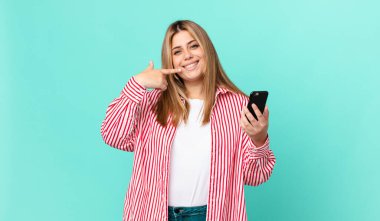 Kıvrımlı, güzel, sarışın kadın kendi gülüşünü işaret ediyor ve akıllı bir telefon tutuyor.