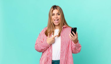 Kıvrımlı, güzel, sarışın kadın heyecanlı ve şaşırmış görünüyor. Yan tarafı gösteriyor ve akıllı bir telefon tutuyor.