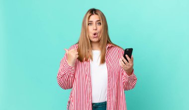 Kıvrımlı, güzel sarışın kadın inançsızlıktan şaşkına dönmüş ve elinde akıllı bir telefon tutuyor.