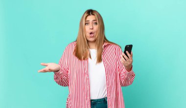 Eğri büğrü, güzel, sarışın kadın inanılmaz bir sürprizle şaşkın ve şaşkın ve akıllı bir telefon tutuyor.