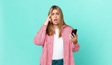 Kıvrımlı, güzel, sarışın kadın şaşırmış görünüyor, yeni bir düşünce, fikir ya da konsept buluyor ve elinde akıllı bir telefon tutuyor.