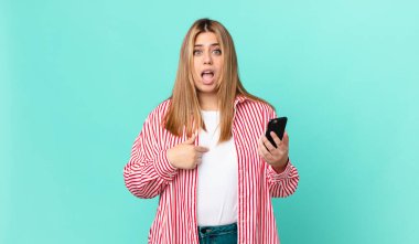 Kıvrımlı, güzel sarışın kadın şok olmuş ve şaşırmış görünüyor ağzı açık, kendini işaret ediyor ve akıllı bir telefon tutuyor.