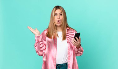 Kıvrımlı, güzel, sarışın kadın şaşırmış ve şok olmuş görünüyor. Çenesi düşmüş, elinde bir nesne, elinde akıllı bir telefon tutuyor.