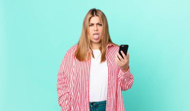 Kıvrımlı, güzel, sarışın bir kadın. Tiksinmiş ve rahatsız olmuş hissediyor. Dilini dışarı çıkarıyor ve akıllı bir telefon tutuyor.