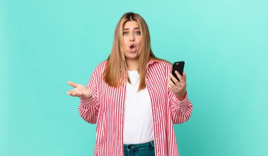 Kıvrımlı, güzel, sarışın kadın şok olmuş, şaşırmış ve elinde akıllı bir telefon tutuyor.