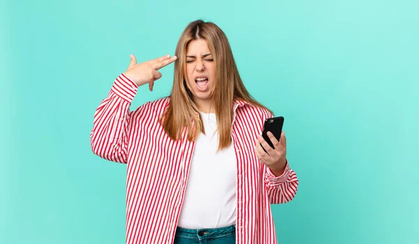Kıvrımlı güzel sarışın kadın mutsuz ve stresli görünüyor, intihar jesti silah işareti yapıyor ve elinde akıllı bir telefon var.