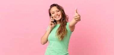 Genç ve güzel bir kadın gururlu, olumlu gülümsüyor, parmakları havada ve akıllı bir telefon tutuyor.