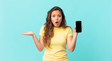 Genç ve güzel bir kadın şaşırmış ve şok olmuş görünüyor, çenesi düşmüş, elinde bir nesne, elinde akıllı bir telefon tutuyor.