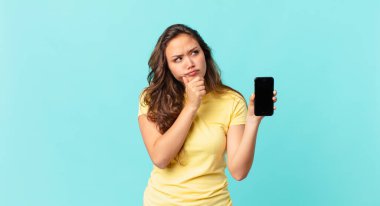 Genç, güzel bir kadın, düşünüyor, kuşkulu ve şaşkın hissediyor ve akıllı bir telefon tutuyor.