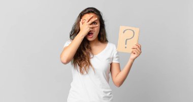 Genç, güzel bir kadın şok olmuş, korkmuş ya da korkmuş görünüyor. Yüzü elle kapatılmış ve elinde bir soru işareti var.