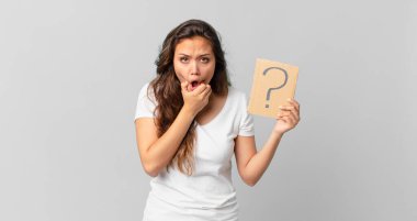 Ağzı ve gözleri açık, eli çenesinde ve elinde soru işareti olan genç ve güzel bir kadın.