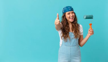 Genç ve güzel bir kadın gururlu, olumlu gülümsüyor, parmakları havada ve bir duvar boyuyor.