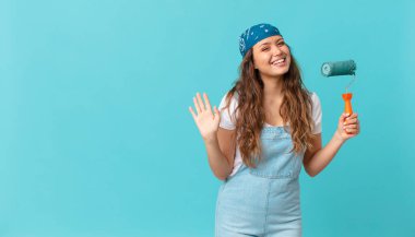 Genç ve güzel bir kadın mutlu bir şekilde gülümsüyor, el sallıyor, seni selamlıyor ve bir duvar boyuyor.