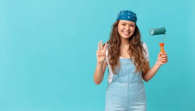 Genç ve güzel bir bayan gülümsüyor ve arkadaş canlısı görünüyor, üç numarayı gösteriyor ve bir duvar boyuyor.