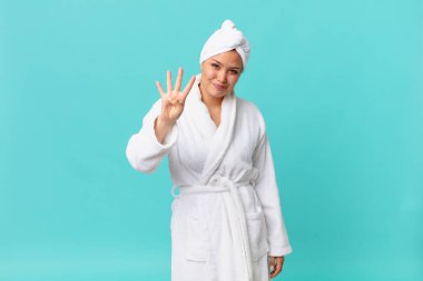 Genç, güzel bir kadın gülümsüyor ve arkadaş canlısı görünüyor. Dört numarayı gösteriyor ve duştan sonra banyo cübbesi giyiyor.