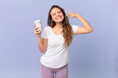 Genç ve güzel bir kadın, kendine güvenerek gülümseyerek kendi gülüşünü gösteriyor ve elinde bir kahve tutuyor.