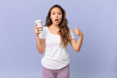 Genç ve güzel bir kadın ağzı açık, kendini gösteren ve elinde kahve tutan şok olmuş ve şaşırmış görünüyor.