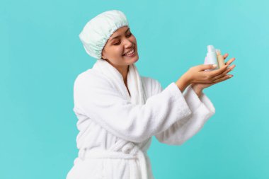 Bornoz giyen ve elinde temiz bir ürün şişesi tutan genç ve güzel bir kadın.