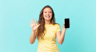 Genç, güzel bir bayan gülümsüyor ve arkadaş canlısı görünüyor. Beş numarayı gösteriyor ve elinde akıllı bir telefon var.