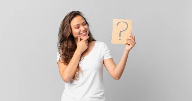 Genç ve güzel bir kadın mutlu bir şekilde gülümser ve hayal kurar ya da şüphe eder ve elinde soru işareti taşır.