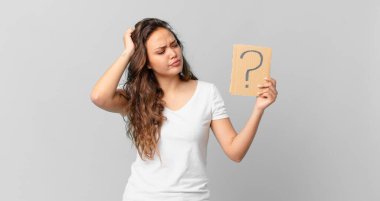 Genç, güzel bir kadın kafası karışmış ve kafası karışmış hissediyor. Kafasını kaşıyor ve elinde soru işareti taşıyor.