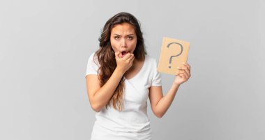 Ağzı ve gözleri açık, eli çenesinde ve elinde soru işareti olan genç ve güzel bir kadın.