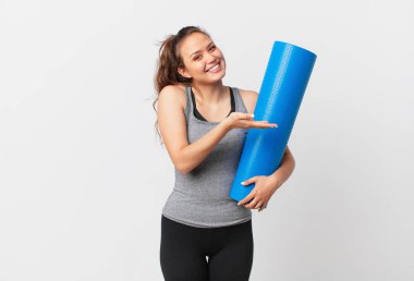 Genç, güzel bir kadın neşeyle gülümsüyor, mutlu hissediyor ve bir konsept gösteriyor ve yoga minderi tutuyor.