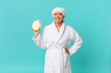 Genç, güzel bir kadın mutlu bir şekilde gülümsüyor, eli kalçasında, kendinden emin ve duştan sonra banyo cübbesi giyiyor.