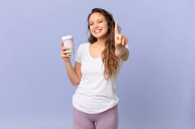 Genç ve güzel bir kadın gururla gülümsüyor ve kendine güvenerek bir numara yapıyor ve elinde kahve tutuyor.