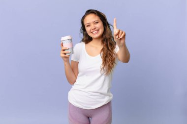 Genç, güzel bir bayan gülümsüyor ve arkadaş canlısı görünüyor. Bir numarayı gösteriyor ve elinde kahve tutuyor.