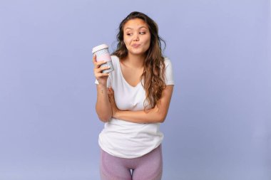 Genç, güzel bir kadın omuz silkiyor, kafası karışık ve kararsız ve elinde kahve tutuyor.