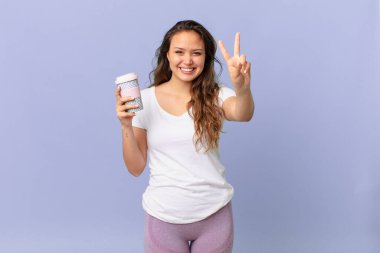 Genç, güzel bir bayan gülümsüyor ve arkadaş canlısı görünüyor. İki numarayı gösteriyor ve elinde kahve tutuyor.