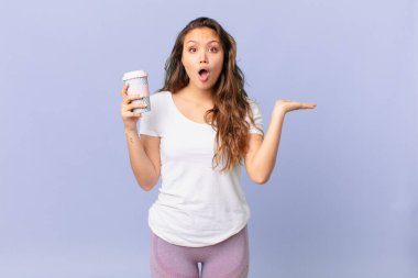 Genç ve güzel bir kadın şaşırmış ve şok olmuş görünüyor, çenesi düşmüş, elinde bir nesne, elinde kahve tutuyor.