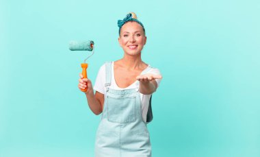 Güzel sarışın kadın mutlu bir şekilde gülümsüyor ve bir konsept sunuyor ve bir duvar boyuyor.