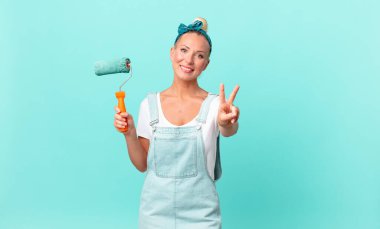 Güzel sarışın kadın gülümsüyor ve mutlu görünüyor, zafer veya barış işareti yapıyor ve bir duvar boyuyor.