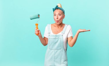 Güzel sarışın kadın şaşırmış ve şok olmuş görünüyor, çenesi düşmüş bir nesneyi tutuyor ve bir duvar boyuyor.